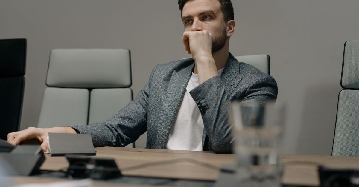 Homem em blazer sentado em uma mesa de reunião, com uma expressão pensativa.