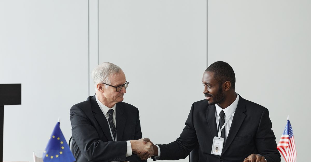 International contracts in brazil inline 1 634393 1786898033 Dois homens em trajes profissionais apertando mãos, com bandeiras europeia e americana ao fundo.