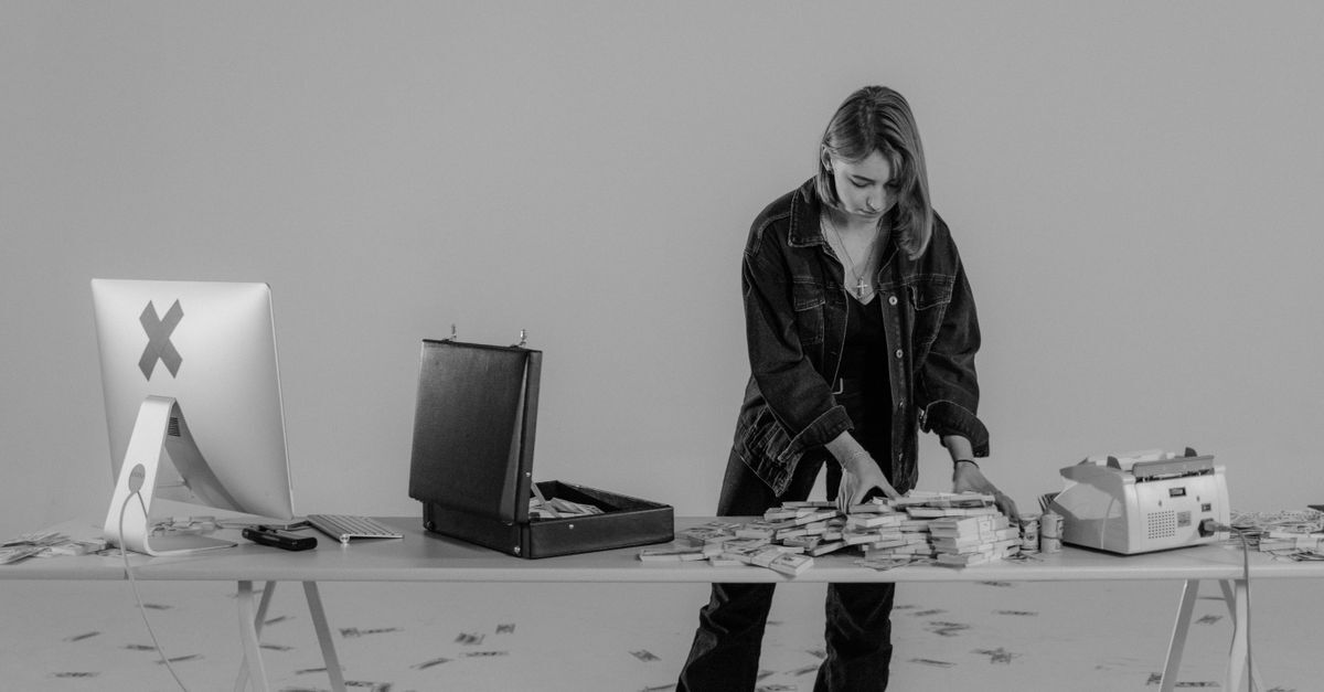 Mulher contando dinheiro em uma mesa com uma caixa de documentos e um computador ao fundo.