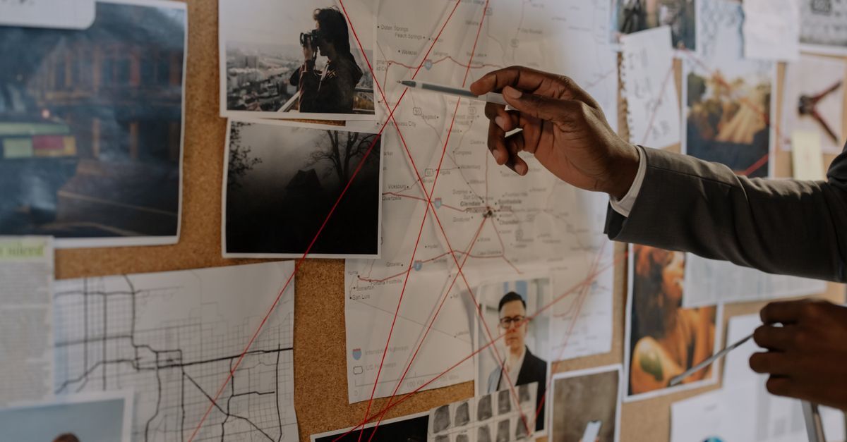 Mão apontando para uma corkboard com fotos e conexões, sugerindo investigação de uma organização criminosa.