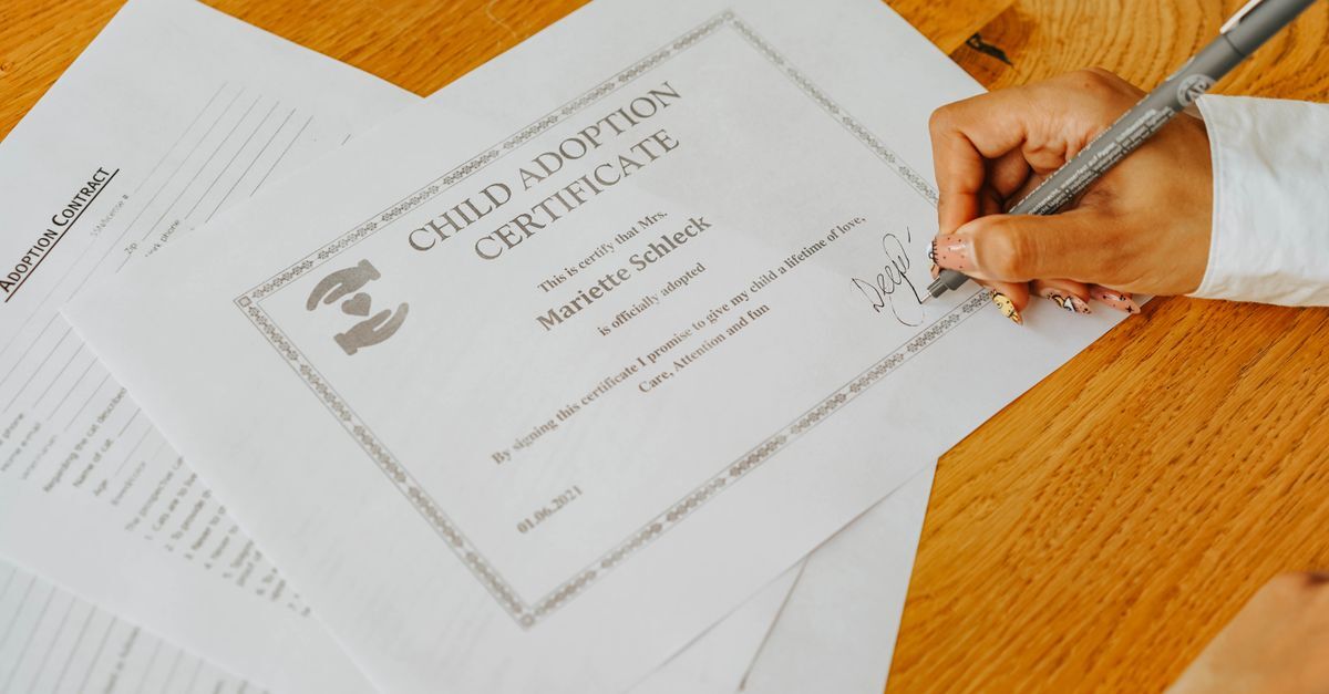 Mão assinando um certificado de adoção de criança em uma mesa de madeira.
